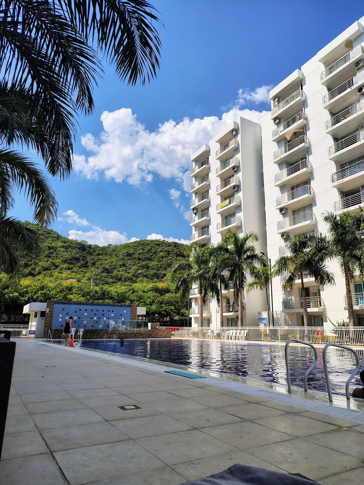 A view of the swimming pool area is captured, showcasing a large pool bordered by sun loungers. Palm trees frame the scene, with a modern building in the background. The clear blue sky features scattered white clouds, enhancing the serene outdoor setting.
