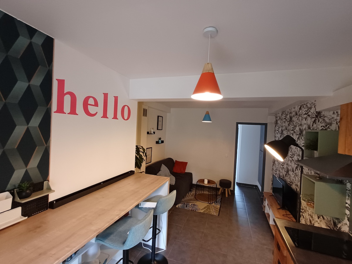 The living area features a modern design with a sectional sofa and a light wood dining table. Two pendant lights of varying colors hang from the ceiling. A decorative wall on the left reads 'hello' in large letters, while the right side showcases a patterned wall, adding character to the space.