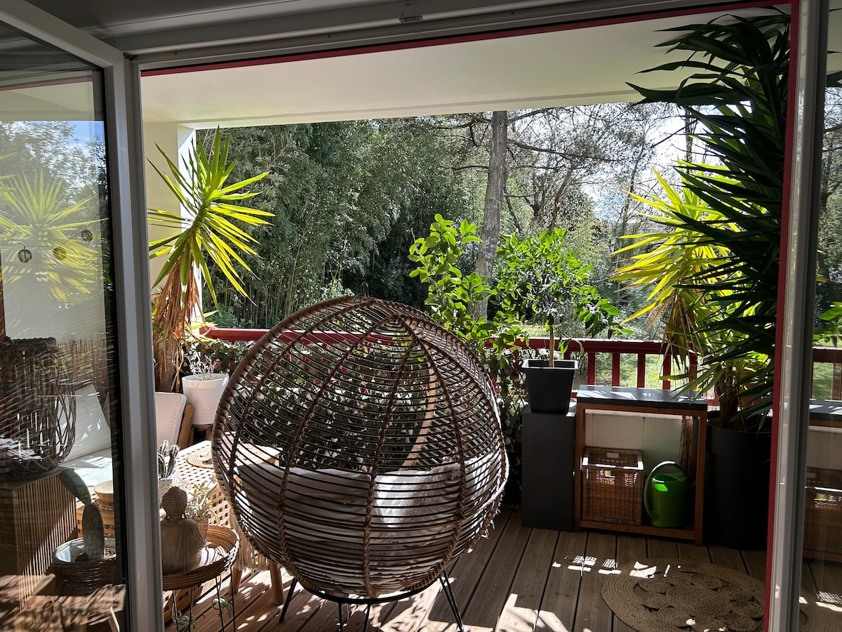 A spacious balcony features an intricate rattan hanging chair positioned among various potted plants. The wooden deck adds warmth, complemented by natural light filtering through the foliage. Greenery from surrounding trees creates a serene backdrop, enhancing the outdoor living space.