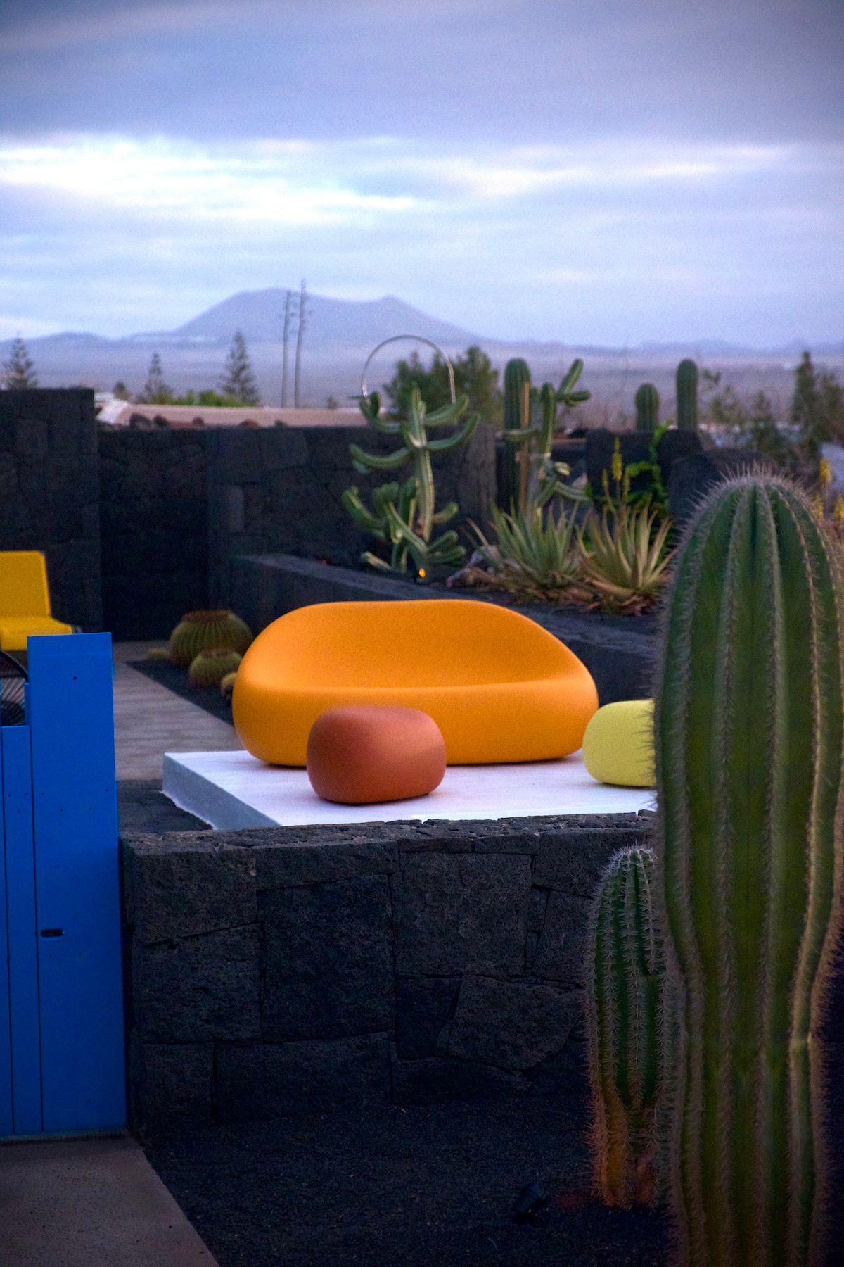 A vibrant outdoor seating area is set against a backdrop of volcanic stone walls and desert plants. Brightly colored furniture, including a round orange chair and assorted small tables, provides a modern touch. Mountains are visible in the distance under a cloudy sky.
