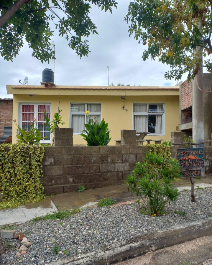 The exterior of a single-story building is presented, featuring a yellow facade with white curtains visible in the window. A low stone wall surrounds the property, with plants and greenery positioned along the front. A sign indicating the name of the department is partially visible.
