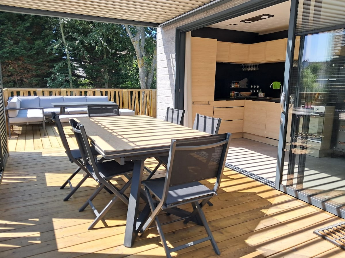 A spacious outdoor terrace features a wooden dining table surrounded by six black chairs. Sunlight filters through the slatted pergola, creating a pattern on the deck. In the background, the kitchen is visible through glass doors, showcasing modern cabinetry and appliances.