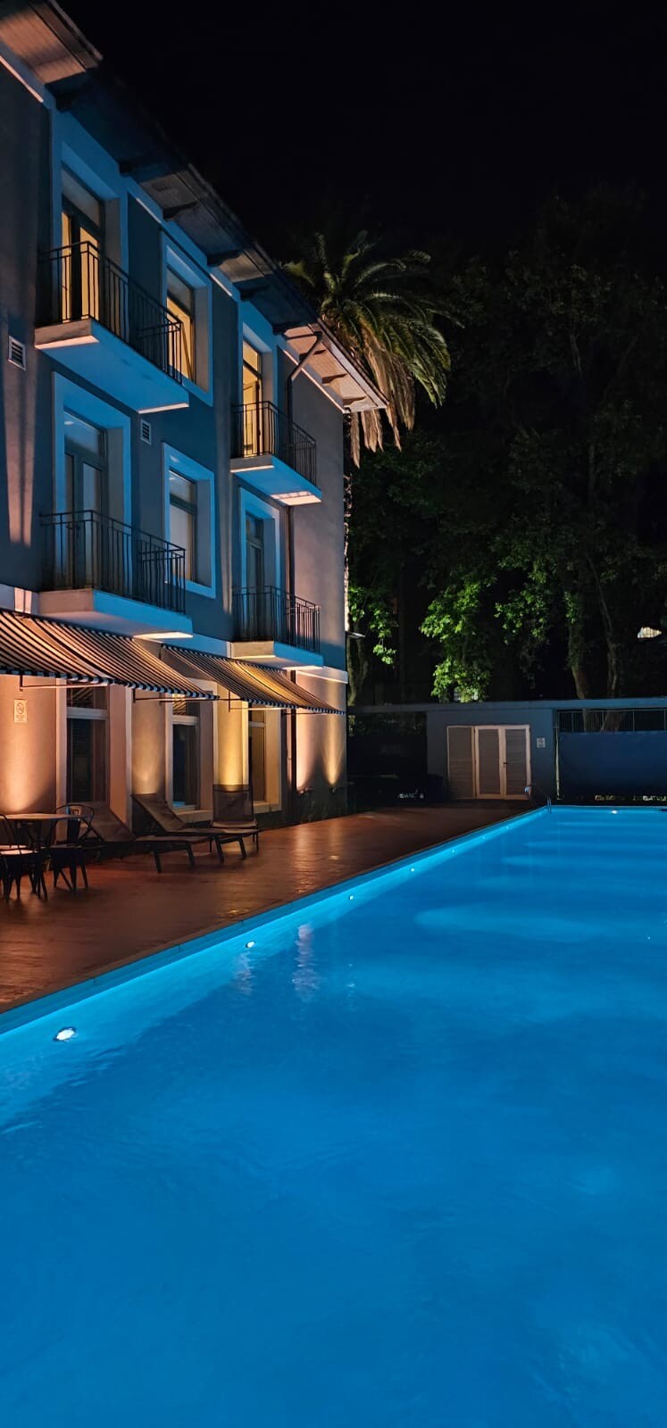 A well-lit pool area features clear blue water, surrounded by a wooden deck. Modern lounge chairs are arranged at the side, while the facade of the building is softly illuminated, showcasing multiple windows and balconies. Lush greenery provides a serene backdrop.