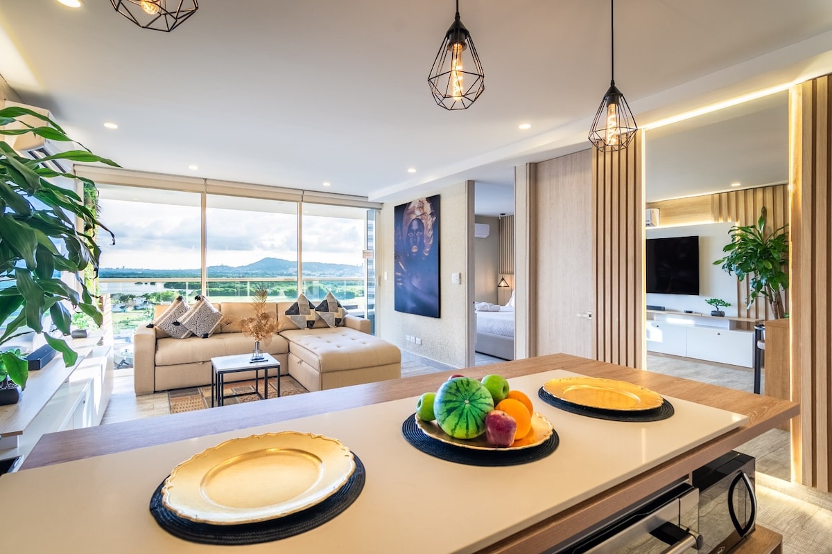 A modern living space features large windows allowing natural light to brighten the area. A comfortable sofa is positioned beside a wooden table, adorned with decorative plates displaying a variety of colorful fruits. Decorative plants enhance the ambiance, creating a serene atmosphere.