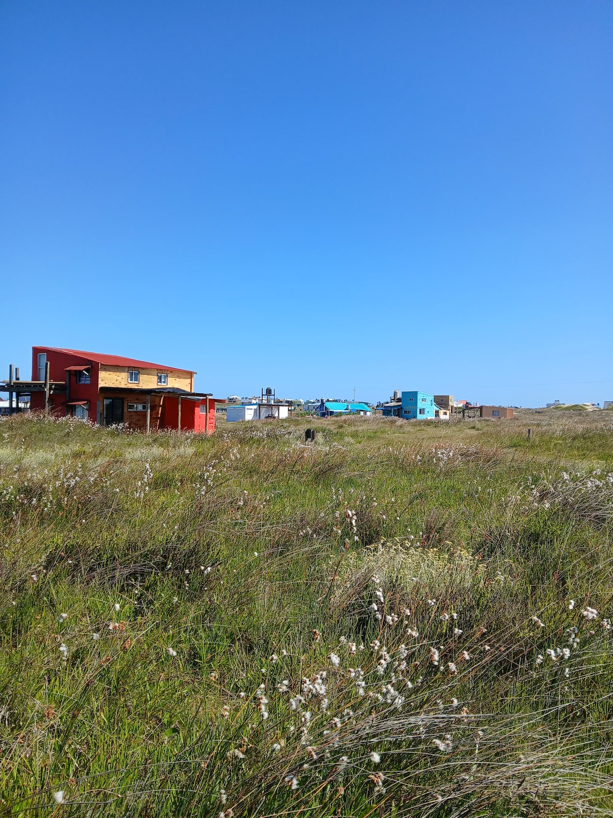 A variety of colorful buildings is visible in the distance, surrounded by tall grasses and wildflowers. The clear blue sky creates a bright backdrop, enhancing the rustic charm of the area. The open landscape suggests a peaceful atmosphere, ideal for relaxation.
