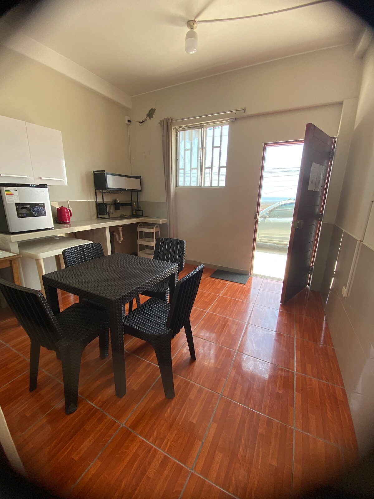A compact kitchen area is visible, featuring a small table with four chairs. Appliances include a microwave, sink, and coffee maker, with bright natural light streaming through the entryway. A door opens to the exterior, enhancing the sense of accessibility.