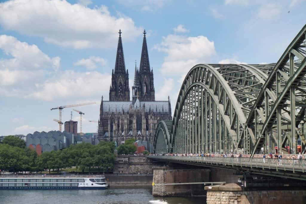 The image captures a view of a river with a large bridge, featuring a distinctive green arch design. In the background, a historic cathedral with two prominent spires towers above the skyline, surrounded by trees and buildings. A boat is visible on the water, adding to the dynamic scene.