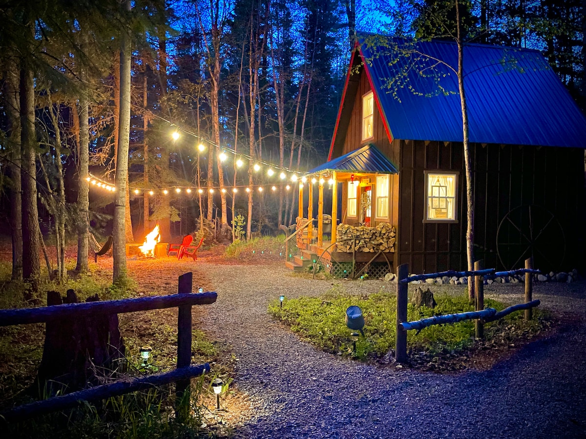 A charming cabin is softly illuminated by string lights in the evening. Red chairs are positioned around a wood-burning fire pit, with a gravel path leading to the covered porch. A stack of firewood is neatly arranged beside the entrance, surrounded by tall trees.