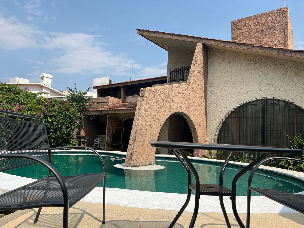 The image captures a unique architectural house with textured walls that complement a serene swimming pool. Black metal chairs and a table are positioned by the poolside, surrounded by lush greenery. The sunny sky provides a bright backdrop to this inviting outdoor space.