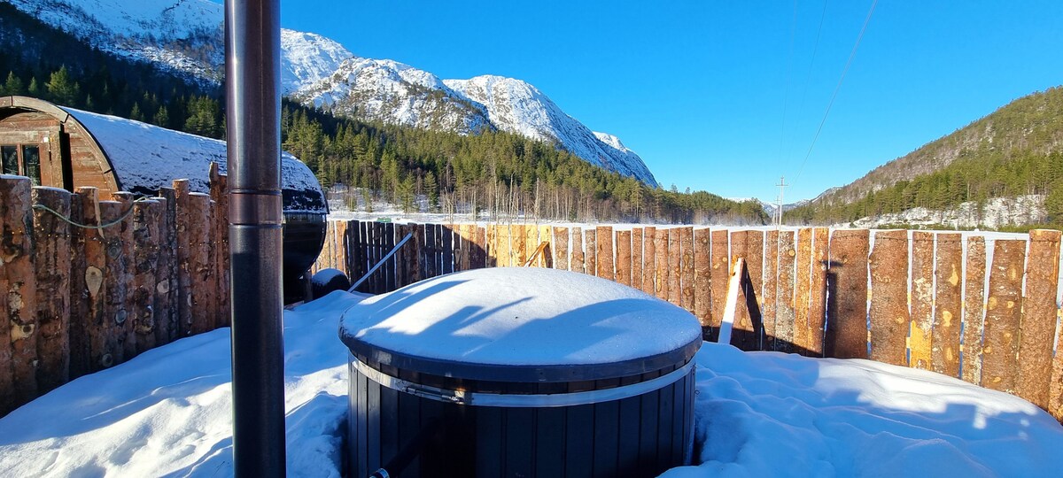 A winter scene featuring a hot tub surrounded by a wooden fence, lightly covered in snow. The distant mountains are visible under a clear blue sky, enhancing the serene ambiance of the rural landscape.