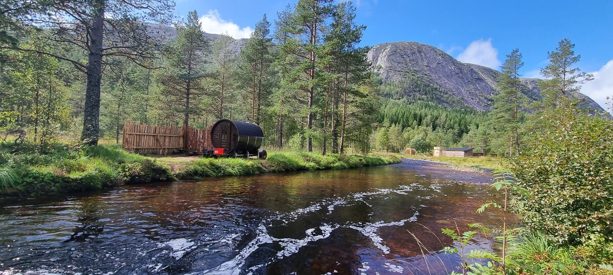 A serene river flows gently through a forested area, bordered by tall trees. A rustic wooden structure sits adjacent to the water, with a cylindrical, dark-colored tub nearby. Rolling green hills and rocky formations create a picturesque backdrop under a clear blue sky.