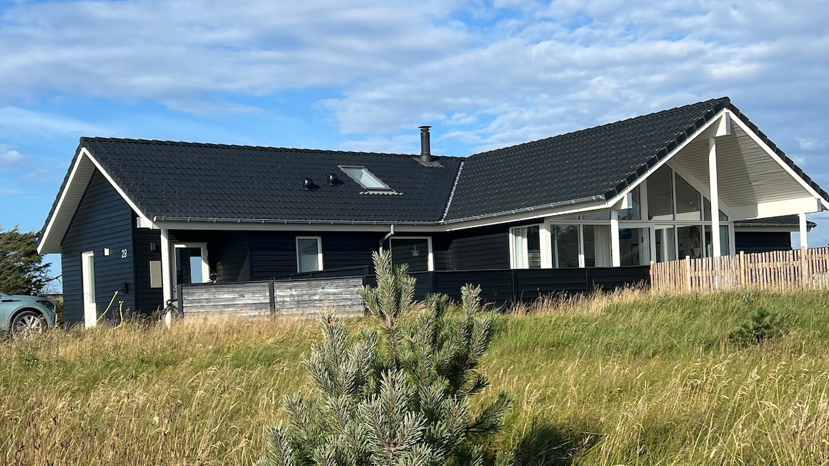 A modern black summer house is shown surrounded by grassy terrain. Large windows allow natural light to fill the spacious interior, while a sloped roof provides architectural interest. A white porch visible at one end offers outdoor seating space, set against a backdrop of subtle hills and a clear sky.
