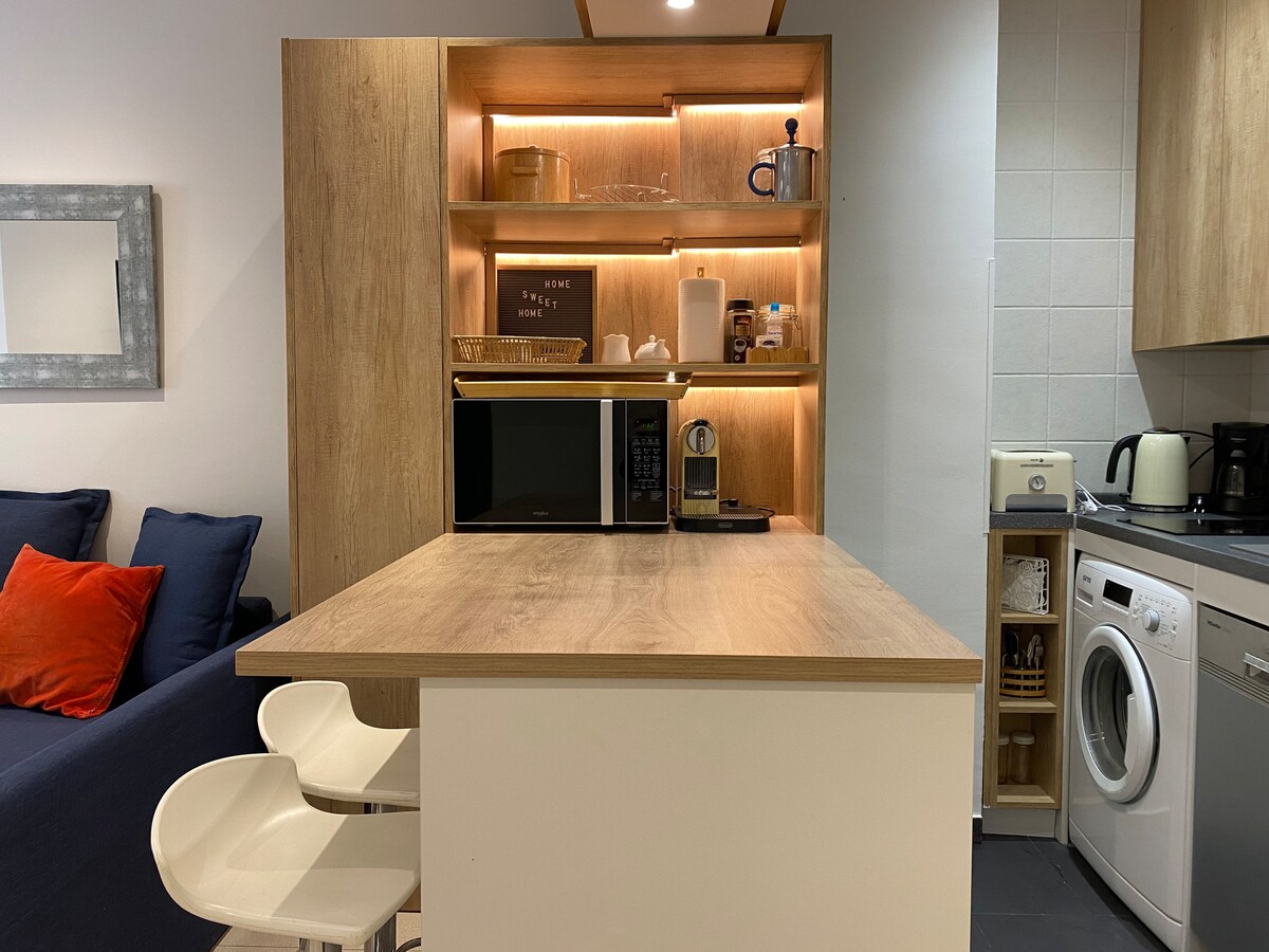 The kitchen area features a wooden countertop with built-in storage above, showcasing neatly arranged items. A microwave, coffee maker, and a washer can be seen nearby. Two white bar stools are positioned in front of the countertop, inviting a casual dining experience.