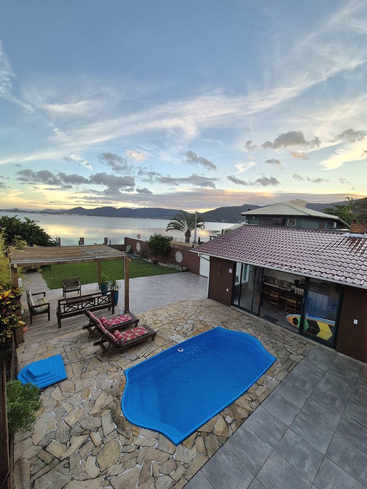 A private outdoor space features a small blue pool, surrounded by stone flooring. A grassy area is visible alongside a shaded pergola with rustic seating. The serene view of the lagoon stretches into the distance, with clouds softly illuminated by the evening light.