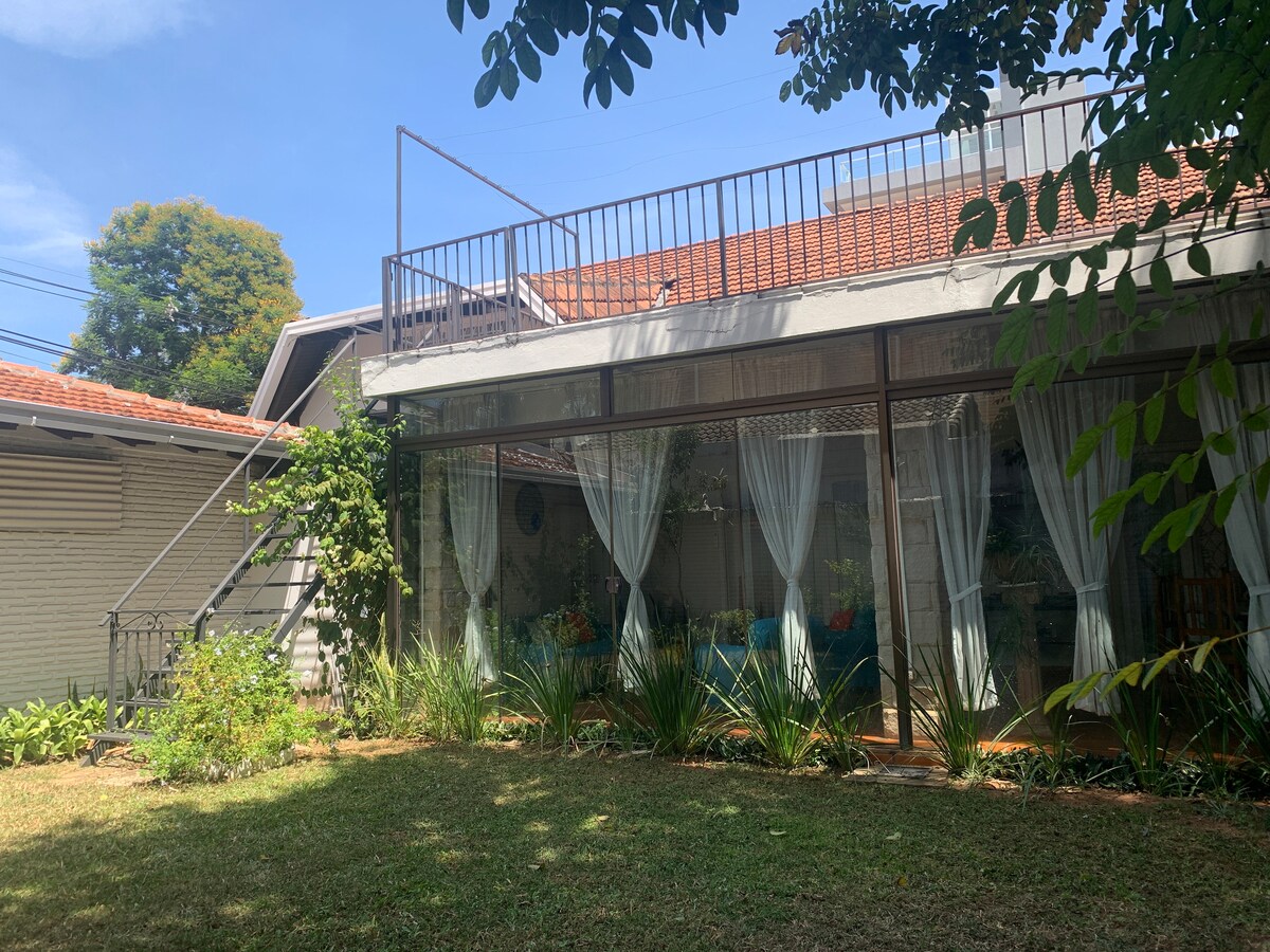 A spacious two-story house is surrounded by greenery, featuring large glass windows adorned with sheer curtains. The upper balcony overlooks the garden, while pathways are lined with plants, adding a natural touch to the exterior.