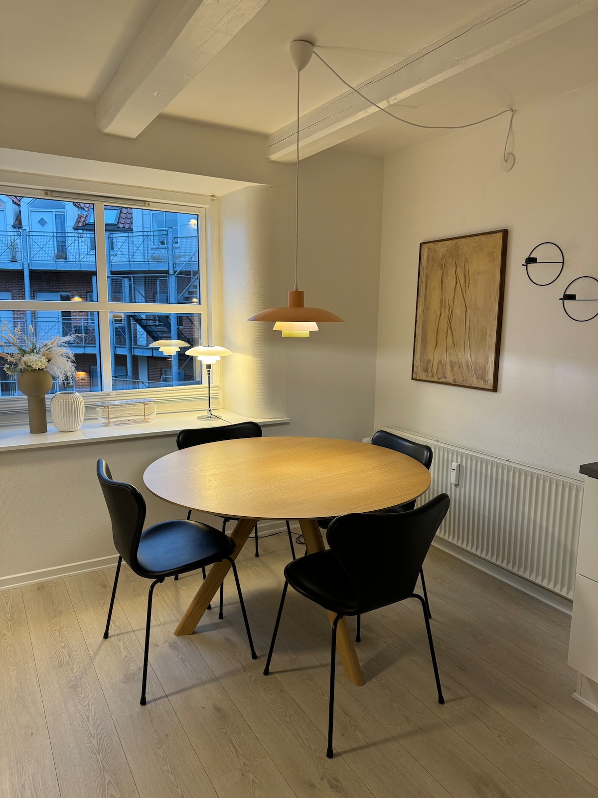 A dining area features a round wooden table surrounded by four black chairs. A pendant light hangs above, illuminating the space. Natural light enters through a window, which provides a view of adjacent buildings. A piece of wall art adds interest to the light-colored walls.