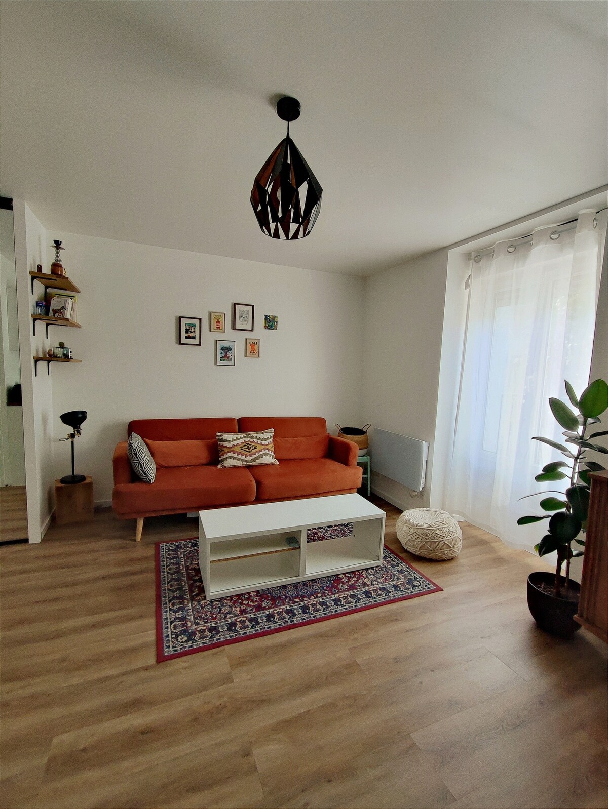 A bright living area is featured, with an orange sofa positioned on a light wood floor. A small coffee table rests on a decorative rug, and a tall plant is placed to the right. Natural light streams in through sheer curtains, enhancing the inviting atmosphere.
