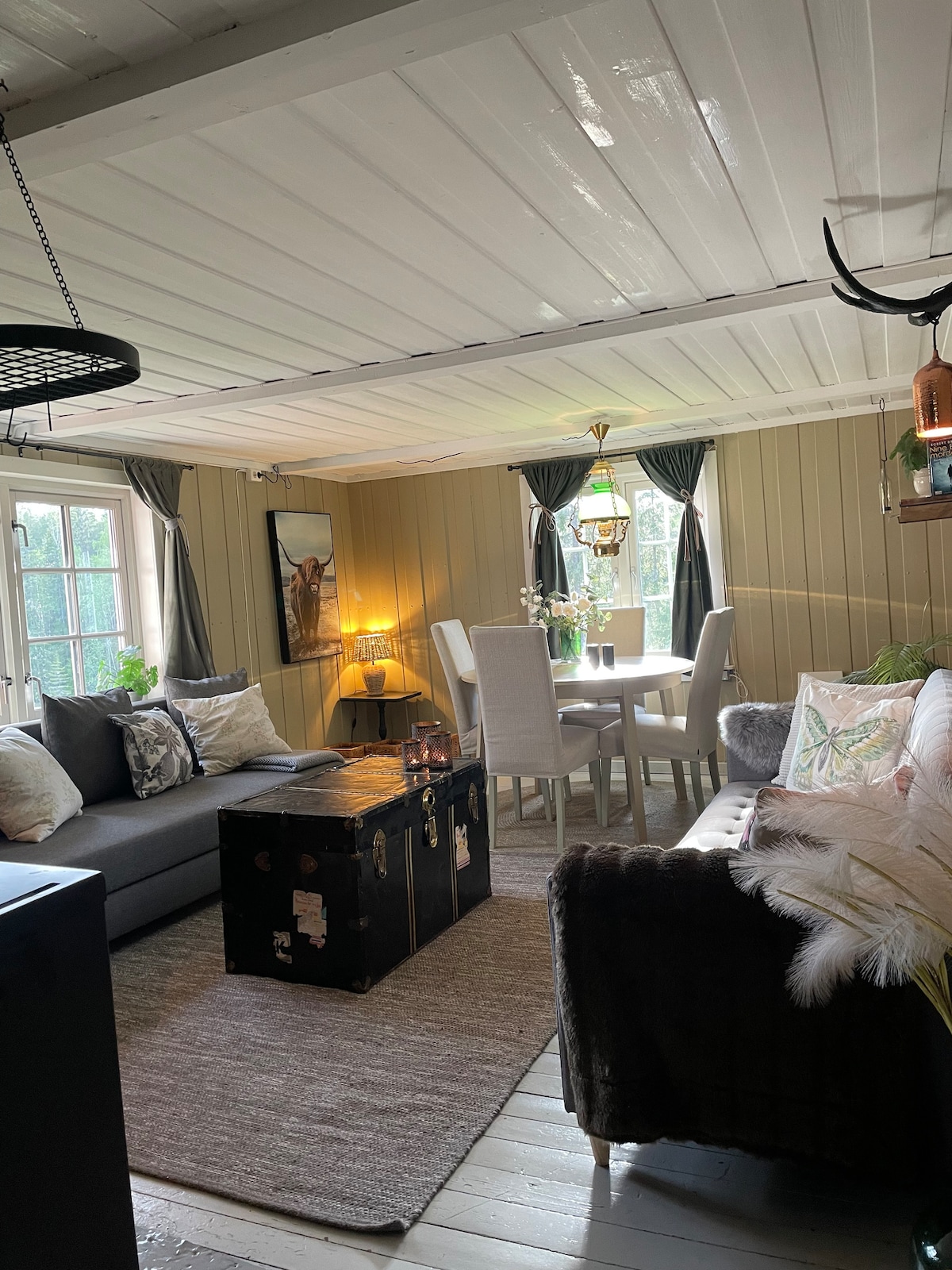 A cozy living area features a dark gray sofa, a vintage trunk, and a round dining table with four white chairs. Light from a decorative lamp highlights the space, complemented by green curtains at the windows. Wooden paneling adds warmth to the ambiance.