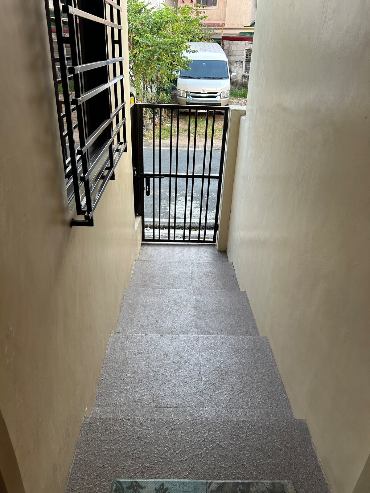 A clean stairway leading to an outdoor gate is visible, with steps finished in neutral tones. A gated entrance is seen at the bottom, and light filters through a nearby window, illuminating the space. A vehicle is parked on the street outside.