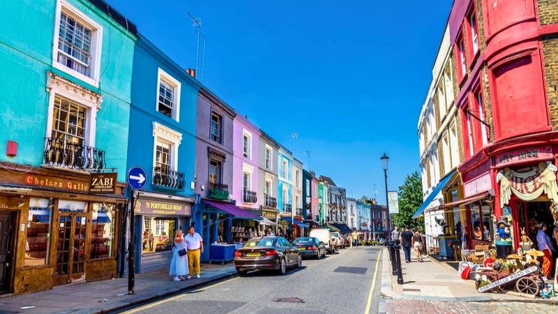 A vibrant street scene showcases colorful buildings with shops and cafes. The architecture reflects a mix of styles, with decorative window balconies and signage. Pedestrians and parked cars add activity to the area under clear blue skies, creating an inviting atmosphere for exploration.