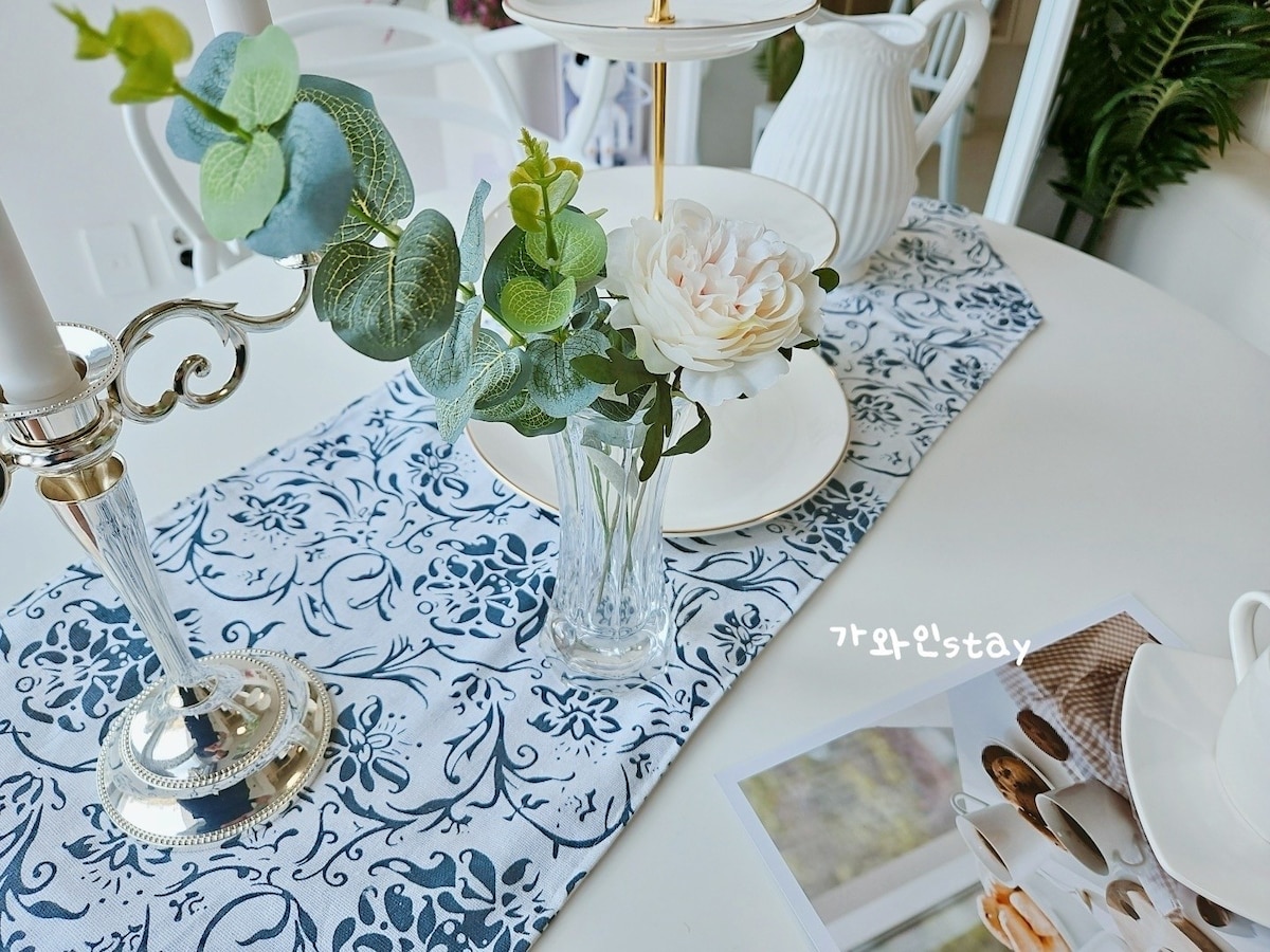A dining table is adorned with an elegant floral arrangement in a clear vase, surrounded by soft greenery and a three-tiered serving stand. A patterned table runner adds a touch of color, complemented by delicate white dinnerware and a thoughtfully placed magazine.