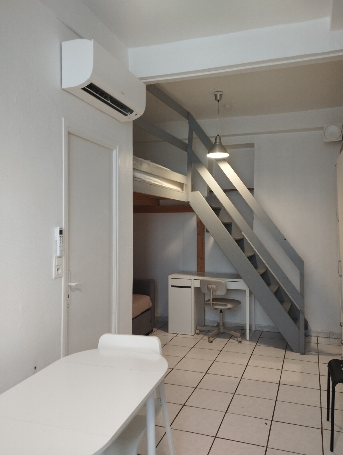 The interior of a studio apartment is showcased, featuring a modern ladder leading to a mezzanine bed. A small dining table is positioned beside a light-colored chair. An air conditioning unit is visible, along with a cozy sofa and a compact desk, enhancing the functional layout.