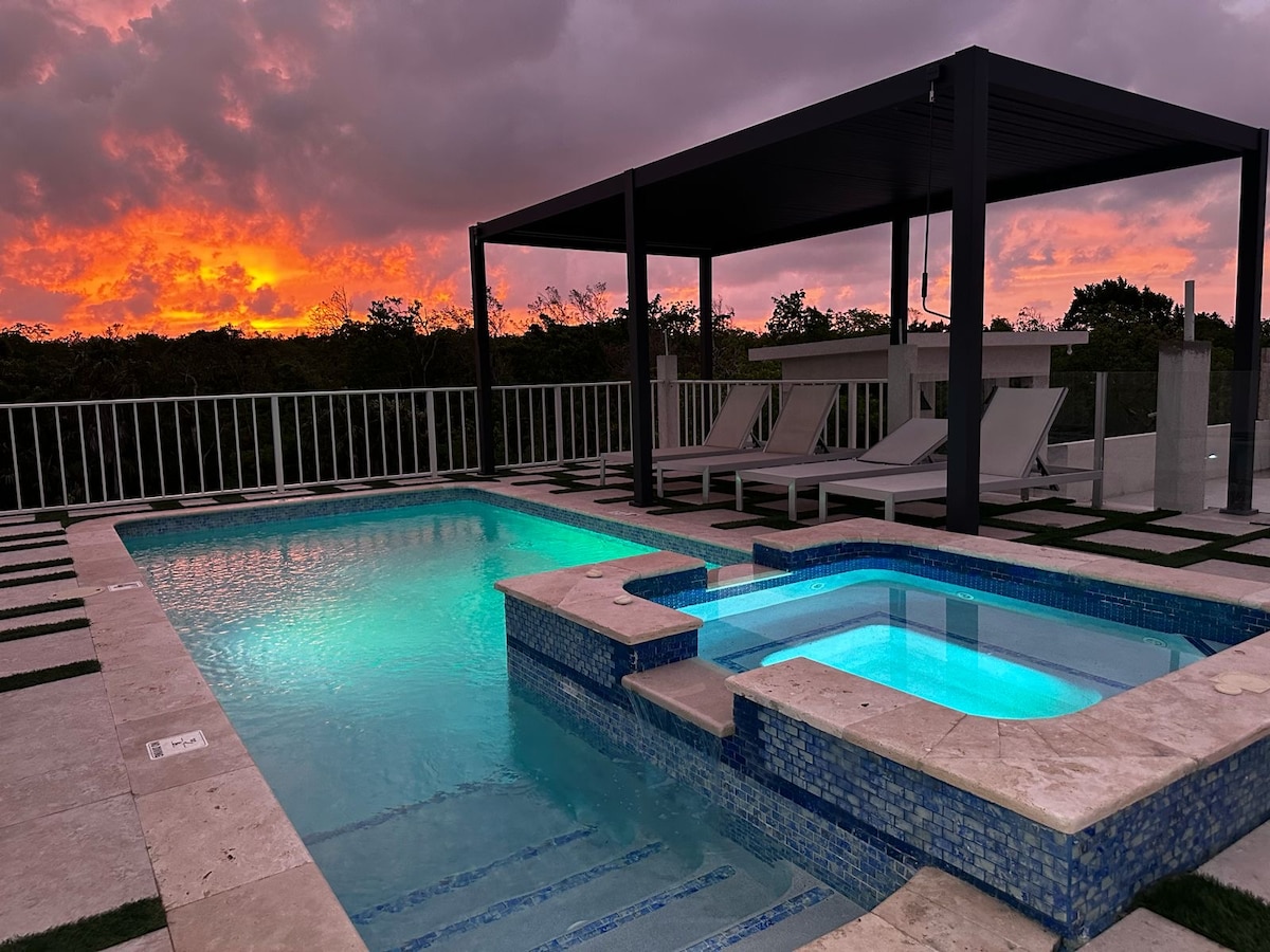 A rooftop pool area is featured, illuminated by soft blue lighting as the sun sets in the background. A covered lounge space is visible, accompanied by two sun loungers. The combination of tiles and grass enhances the outdoor space's design.