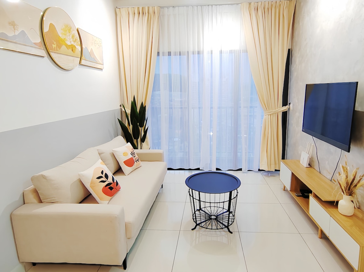 A bright living area features a light-colored sofa adorned with decorative pillows. A small round table sits in front of the couch, and a wall-mounted television is positioned nearby. Large windows with sheer curtains allow natural light to fill the space, creating an open and airy ambiance.