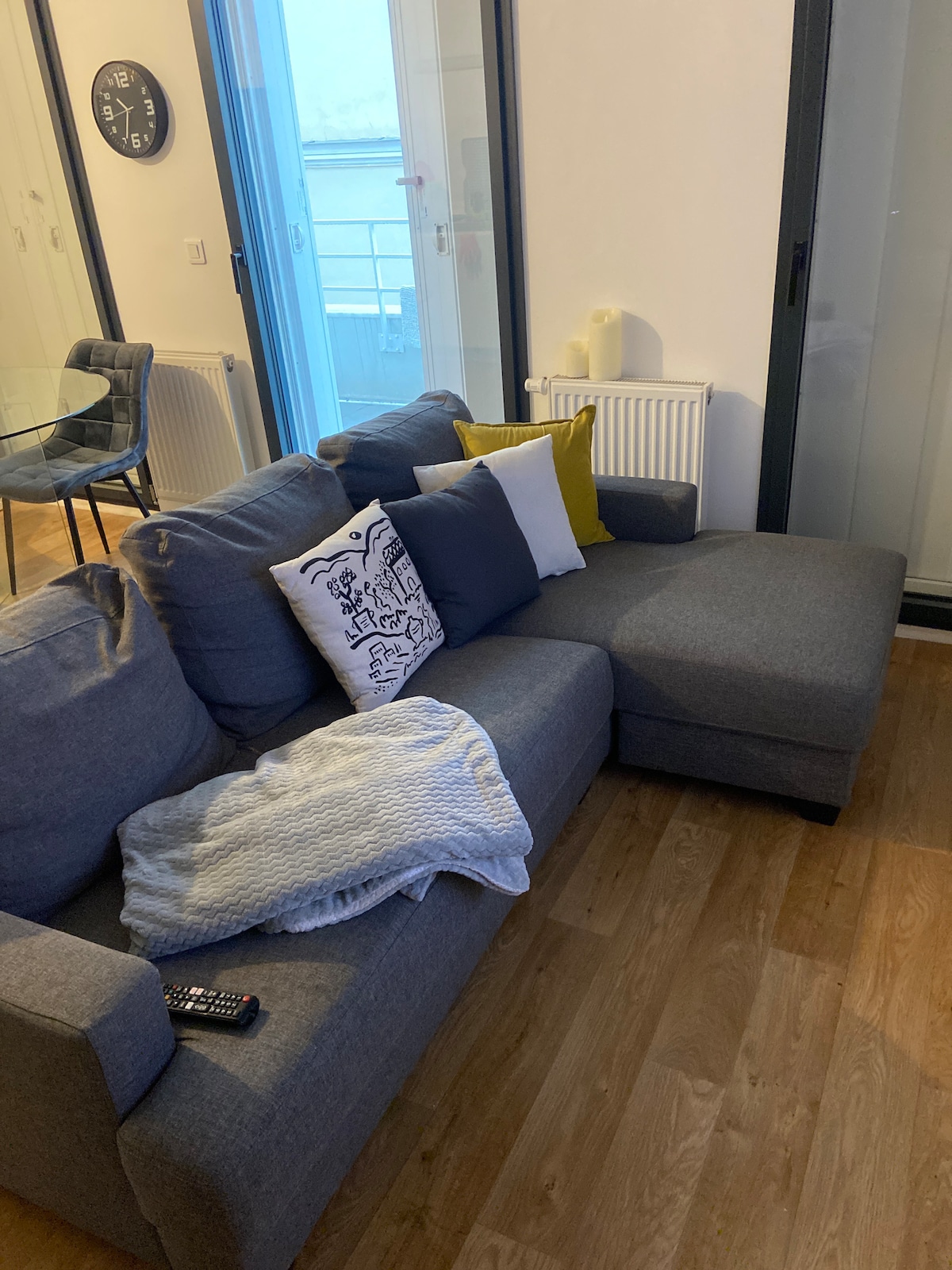 A grey sectional sofa is positioned in the living area, complemented by a mix of decorative pillows in various colors. A light blanket is casually draped over the arm, and a remote control is placed on the cushion. Nearby, a round glass dining table can be seen.
