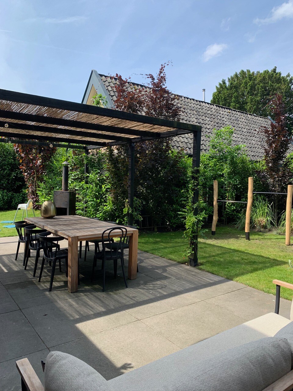 An outdoor patio is showcased, featuring a spacious wooden dining table with black chairs under a pergola. Surrounding greenery provides a natural backdrop, while a lounging area with soft seating is visible. The space is well-lit by natural sunlight.