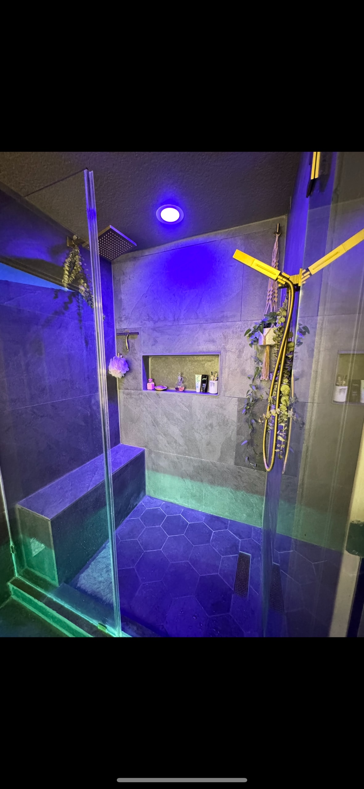 A modern shower area is enclosed by clear glass panels, featuring a built-in bench and a recessed shelf for toiletries. The hexagonal tiled floor is illuminated by dynamic purple and green lighting, enhancing the contemporary design of the space.