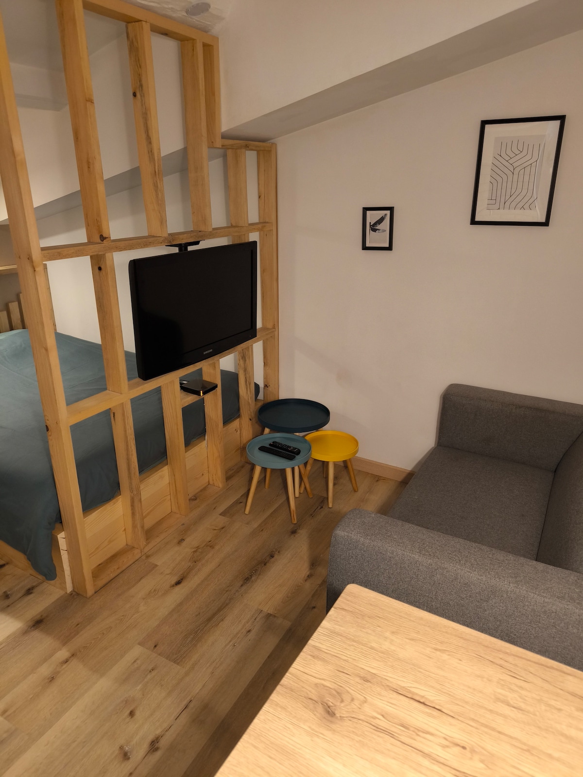 An open space is divided by a wooden frame, creating a cozy sleeping area with a bed behind it. A flat-screen TV is mounted on the frame, while a small side table with two colorful stools and a gray sofa occupy the foreground. Minimalist wall art complements the design.