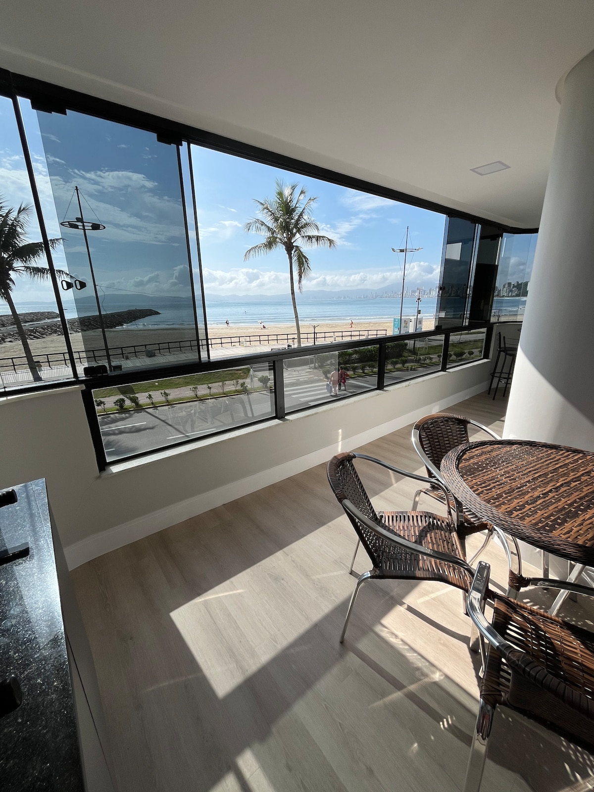A dining area is presented with a round table and four wicker chairs, positioned near large glass windows that overlook palm trees and the ocean. Natural light floods the space, illuminating the modern decor and inviting outdoor scenery.