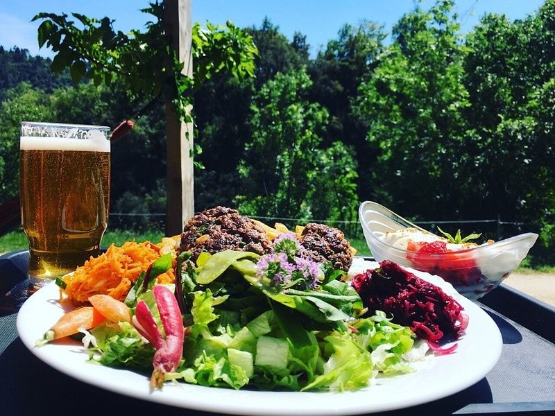 A plate of fresh, colorful salad sits on a table, complemented by two generously sized patties and assorted sides including shredded carrots and beetroot. A glass of beer rests beside the plate, with greenery and trees visible in the background under a clear blue sky.