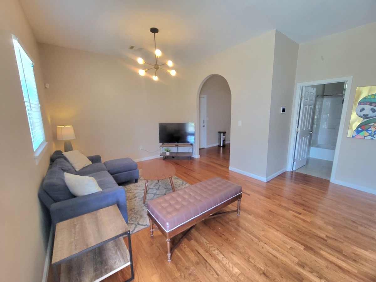 A well-lit living room features a comfortable sofa and a coffee table blending with hardwood flooring. A television is positioned against the wall, with an nearby archway leading to other areas. The space is complemented by a modern light fixture and colorful wall art.