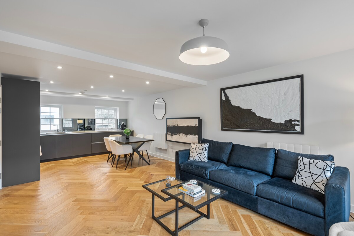 A modern living area features a spacious blue sofa adorned with patterned cushions. A glass coffee table sits in front, with magazines arranged atop. The open-plan design leads to a sleek kitchen area, complemented by a dining table and chairs, all illuminated by natural light.