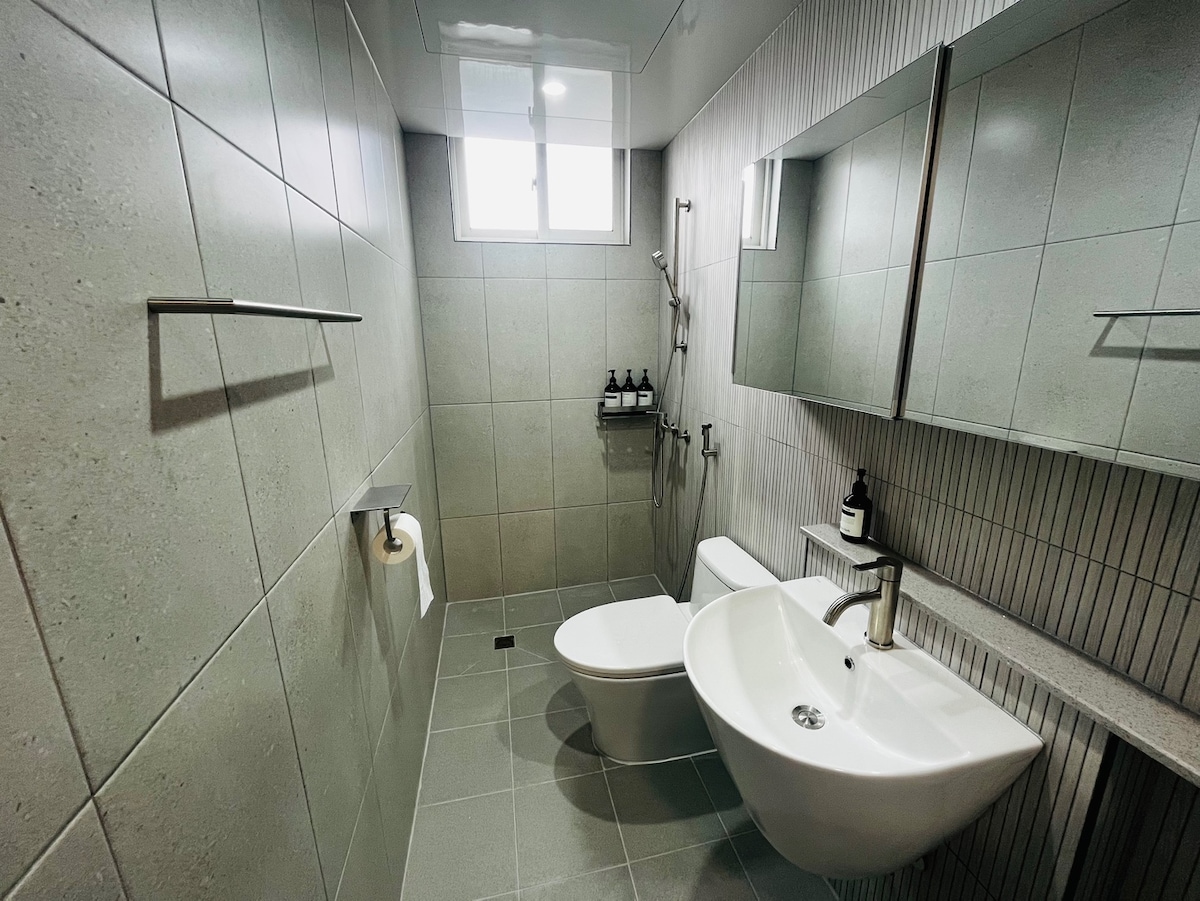 A modern bathroom features a sleek design with large gray tiles covering the walls and floor. A spacious countertop with a stylish sink is accompanied by a wall-mounted mirror. A window allows natural light to illuminate the space, while toiletries are neatly arranged on a shelf.