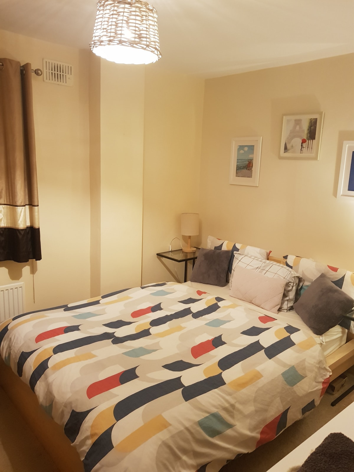 A cozy double bedroom features a large bed adorned with a patterned duvet and assorted pillows. Bedside tables with lamps are positioned on either side, while framed artwork adds a decorative touch to the soft-colored walls.