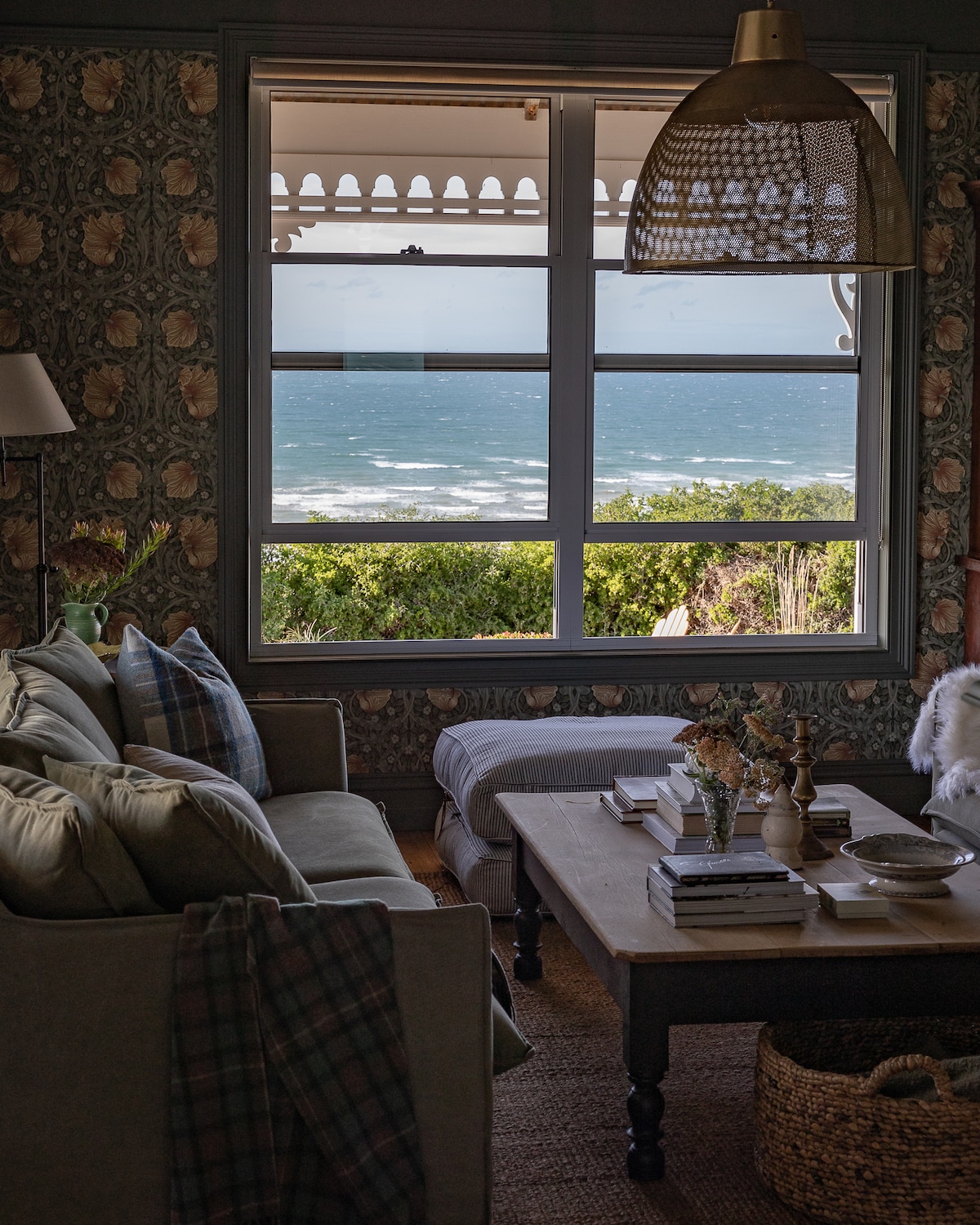 A cozy lounge area is featured, showcasing a comfortable couch adorned with plaid cushions. A wooden coffee table sits in the center, displaying books and decorative vases. Large windows offer a view of the ocean, framed by patterned wallpaper and soft natural light.