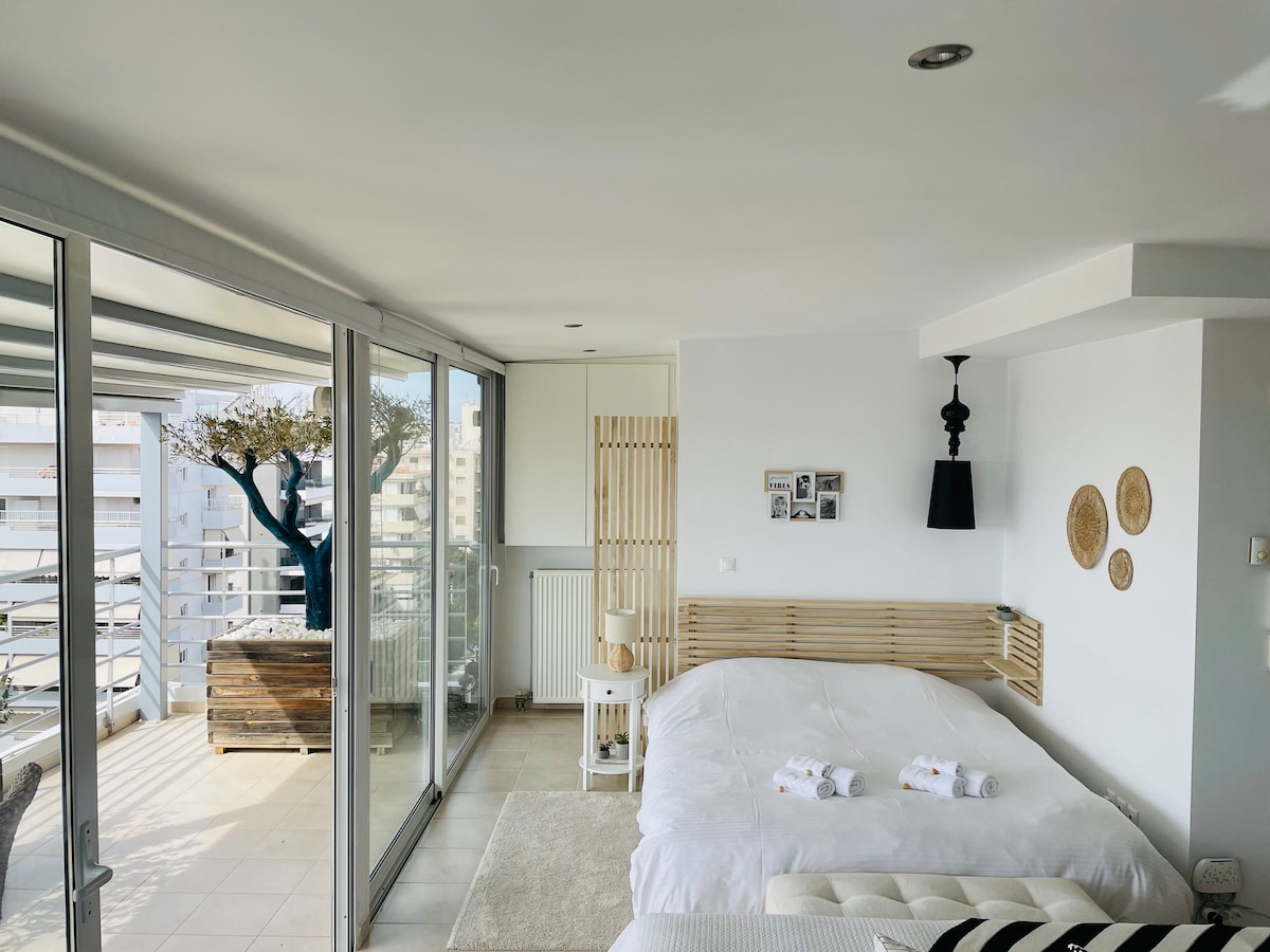 A spacious studio is illuminated by natural light from large glass doors that open to a private balcony. A comfortable bed is arranged with fresh towels, while neutral decor and minimalist furnishings create a serene atmosphere. The balcony features a small table and a tree.