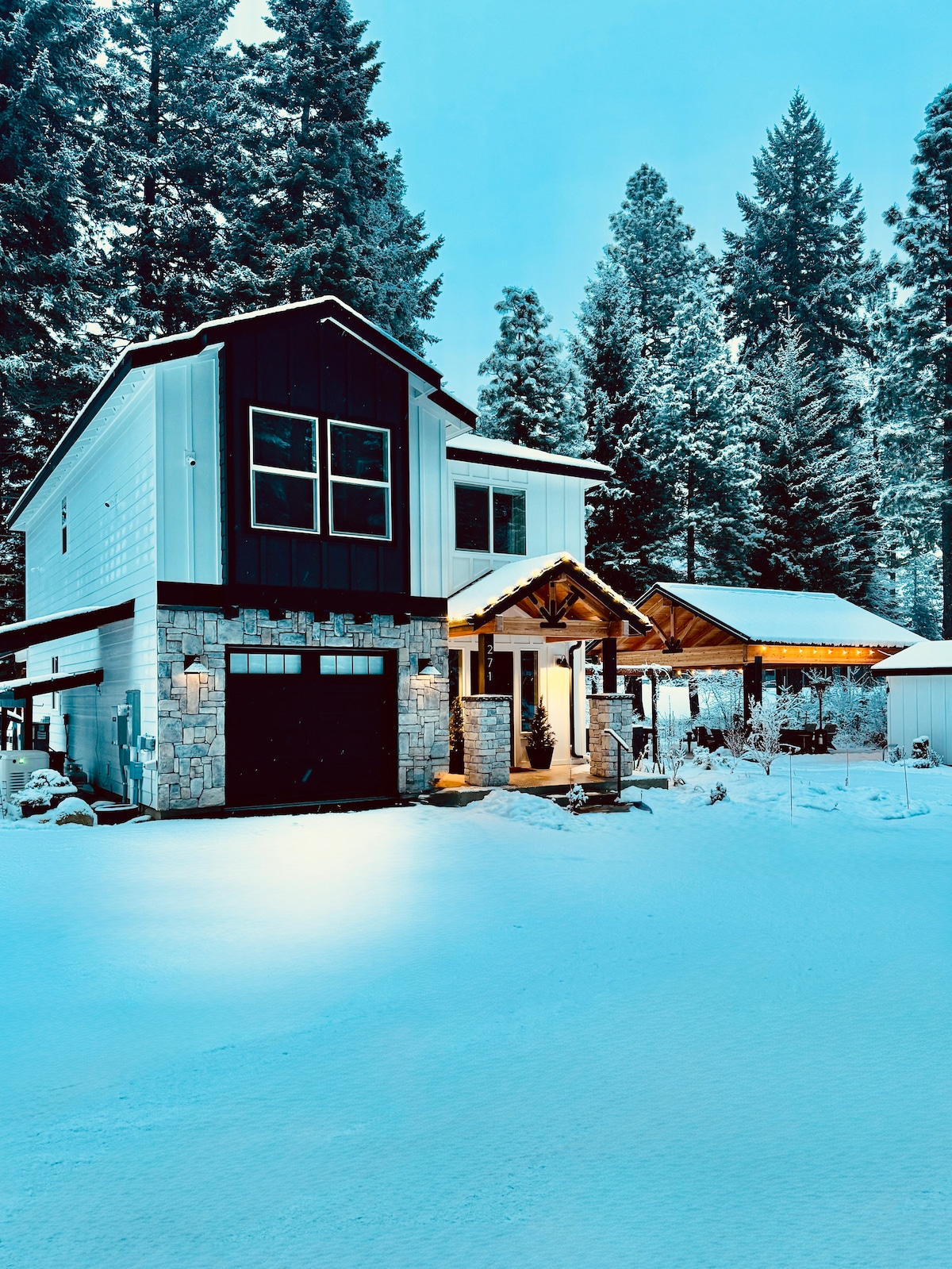A modern two-story farmhouse is set against a snowy landscape, featuring a stone and wood exterior. Soft lights illuminate the entrance, highlighting the inviting overhang and garage. Tall evergreen trees surround the property, enhancing the peaceful winter ambiance.