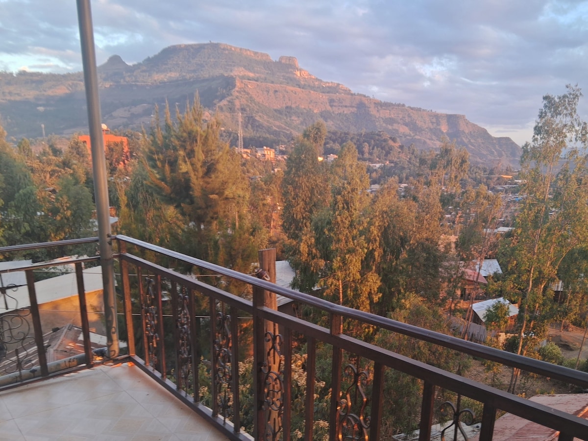 A balcony view captures rolling hills and lush greenery, with the silhouette of a mountain in the background. Sunlight filters through scattered clouds, casting a warm glow over the landscape, which includes a blend of trees and buildings below.