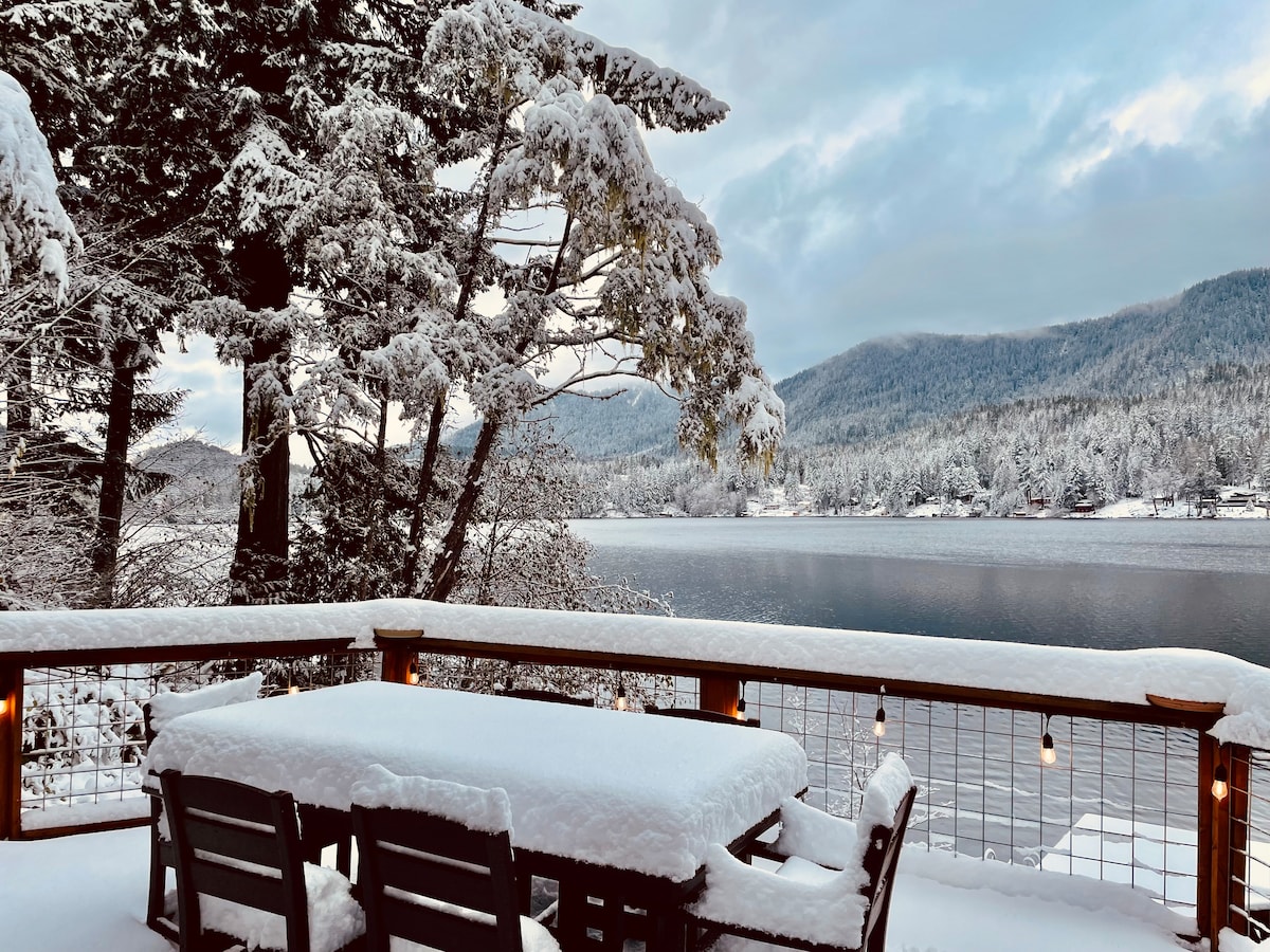 A snow-covered deck overlooks Lake Sutherland, with a table surrounded by chairs blanketed in white snow. Majestic mountains rise in the background, framed by tall evergreens. Soft morning light reflects off the lake's surface, creating a serene winter atmosphere.