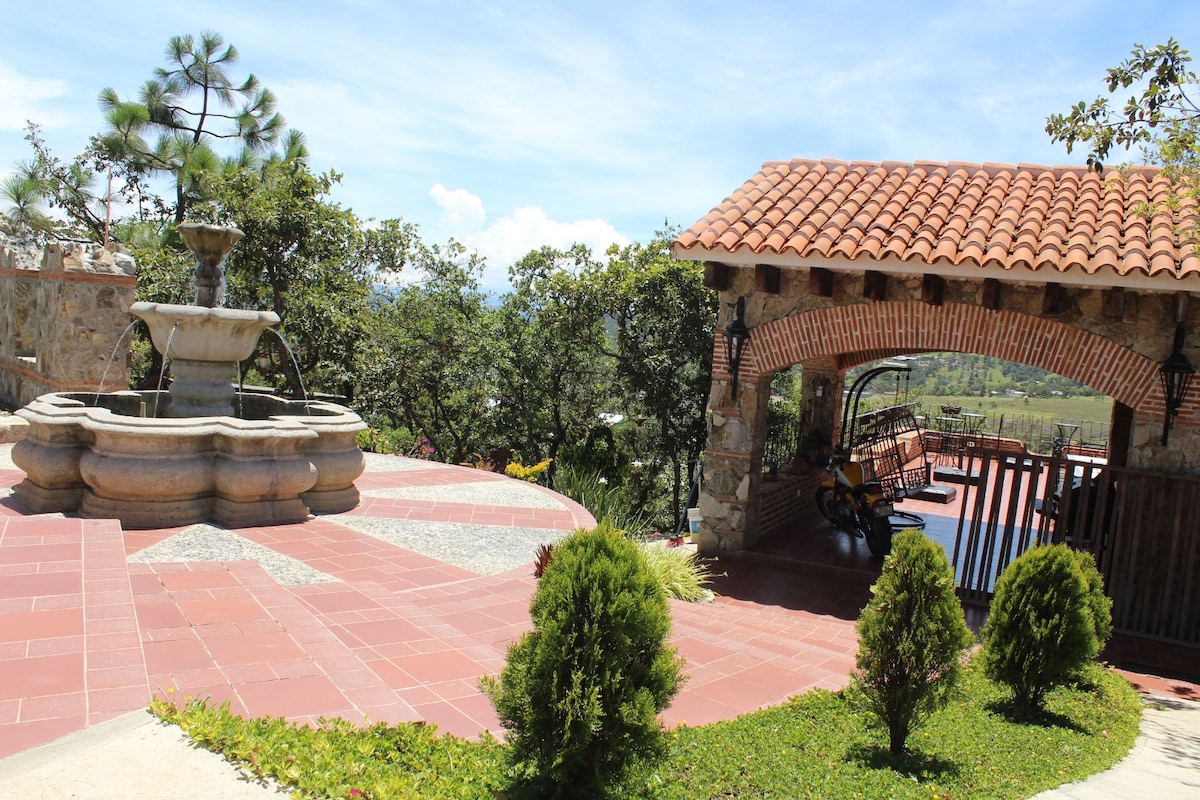 A charming outdoor space features a stone fountain at the center, surrounded by tiled pathways and green landscaping. An open structure with a tiled roof offers seating, with swings visible in the background, surrounded by lush trees and scenic views.