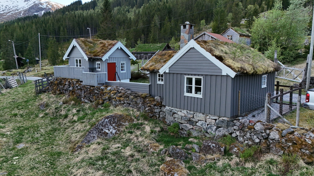 Two small gray cabins with grass roofs are set within a landscaped area. A stone wall surrounds them, providing a rustic charm. Surrounding greenery enhances the natural setting, with mountains visible in the distance.