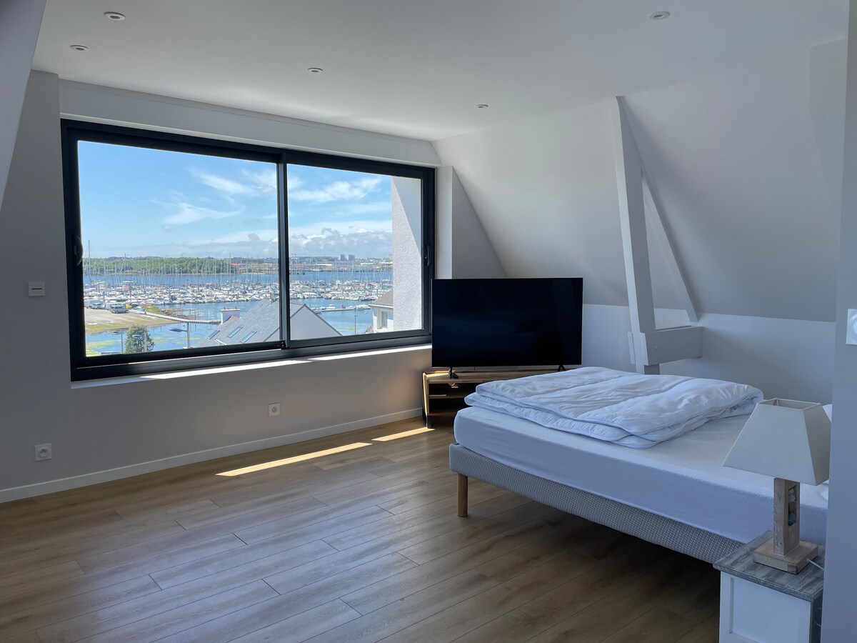 A spacious bedroom is illuminated by natural light flowing through a large window that offers a view of the port. A flat-screen TV is positioned opposite a minimalistic bed, which is partially arranged with bedding. Light wood flooring adds warmth to the modern decor.
