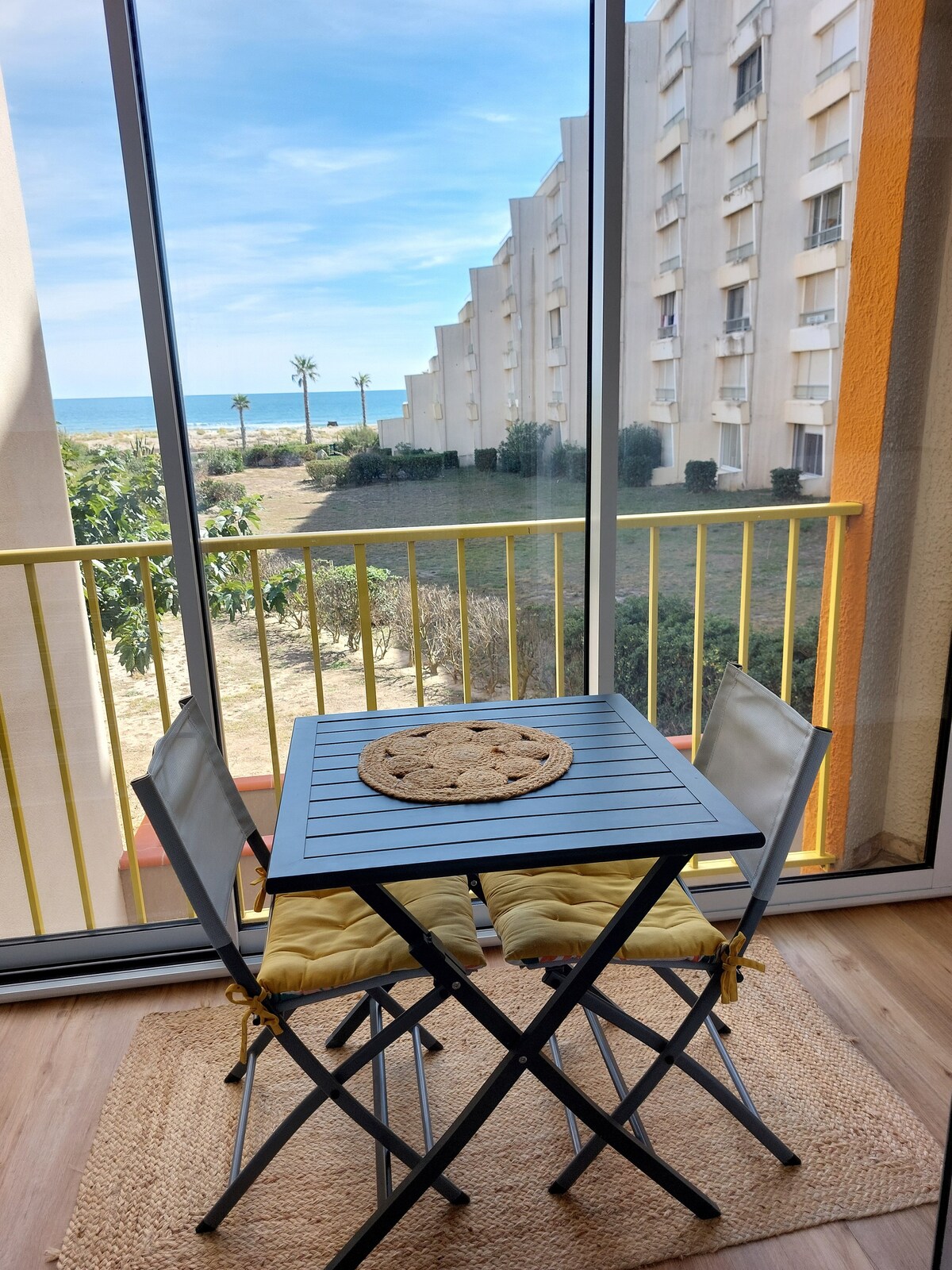 A small table with two chairs is situated in the loggia, providing a view of palm trees and the beach beyond. The space is enclosed with large glass doors, allowing natural light to fill the area, while the ocean can be seen on the horizon.