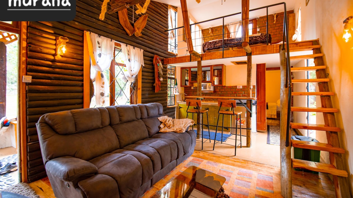 A spacious living area is highlighted by a comfortable gray sofa, with logs and rustic accents adorning the walls. A dining area is visible, alongside a kitchen with wooden details. A staircase leads to a lofted sleeping space, enhancing verticality within the charming interior.