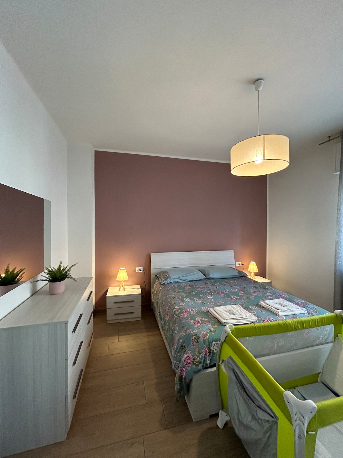 The bedroom features a spacious bed with a floral-patterned bedspread, accompanied by two bedside tables with soft lighting. A light-colored dresser is positioned against one wall, while a bright green crib is placed beside the bed, creating a welcoming family-friendly environment.