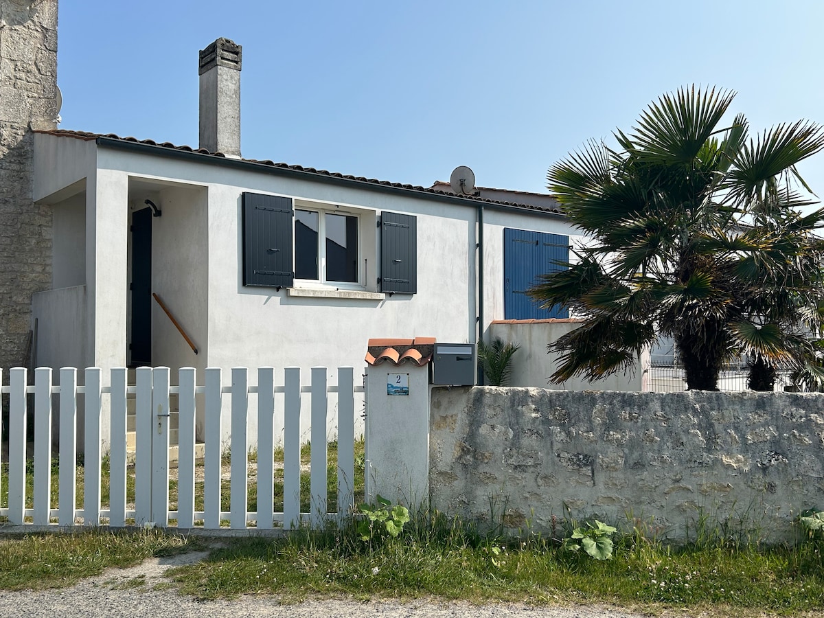 A charming house is visible, featuring a white exterior with shuttered windows and a small staircase leading to the entrance. A palm tree adds a touch of greenery alongside a low stone wall and a wooden fence, creating a welcoming appearance.